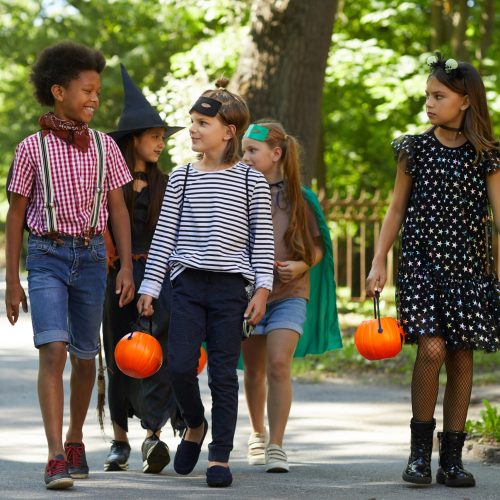 Group of children in costumes with toy pumpkins walking along the street to celebrate Halloween holiday